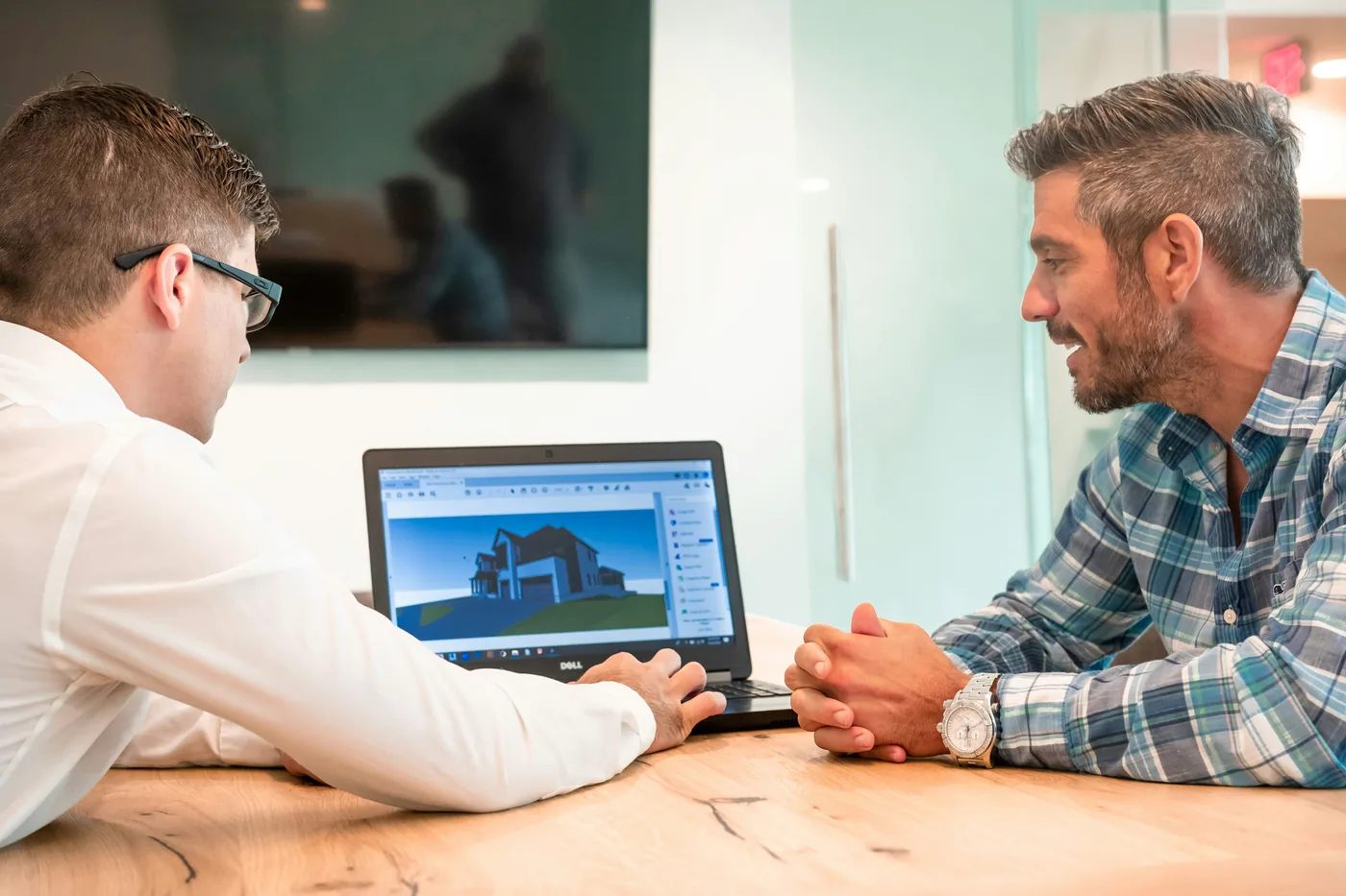 Deux hommes assis à une table devant un ordinateur portable qui affiche un modèle 3D de maison