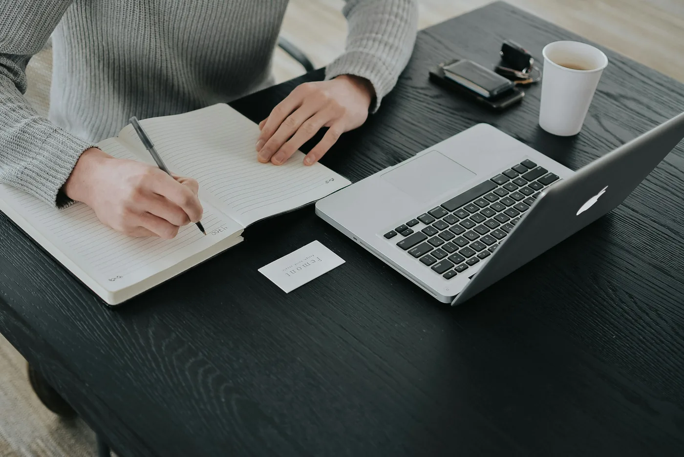Personne qui prend des notes dans un carnet, à côté d'un ordinateur portable, sur un bureau noir