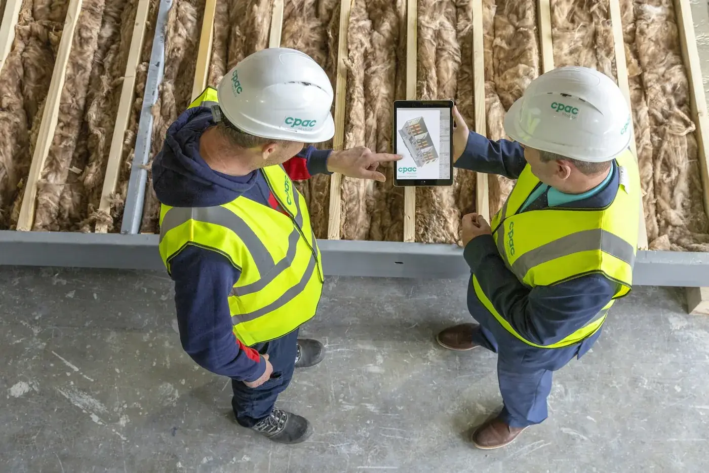 Deux ouvriers en casque et gilet jaune regardent une maquette 3D de bâtiment sur une tablette, sur un plancher en cours de pose